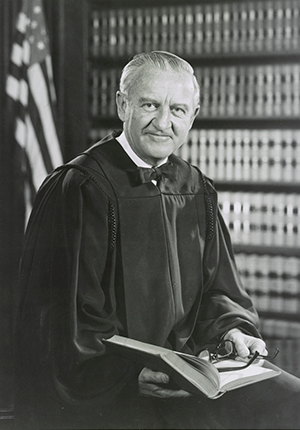 John Paul Stevens, III, three-quarters portrait holding book and wearing judicial robes, seated in front of bookcase and United States flag, by Robert S. Oakes, photographer.